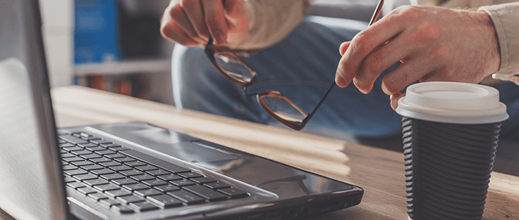 Man putting on glasses in front of a laptop with a travel coffee cup next to it