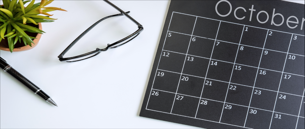 Black and white October calendar sitting on desk next to glasses, a plant, and a pen