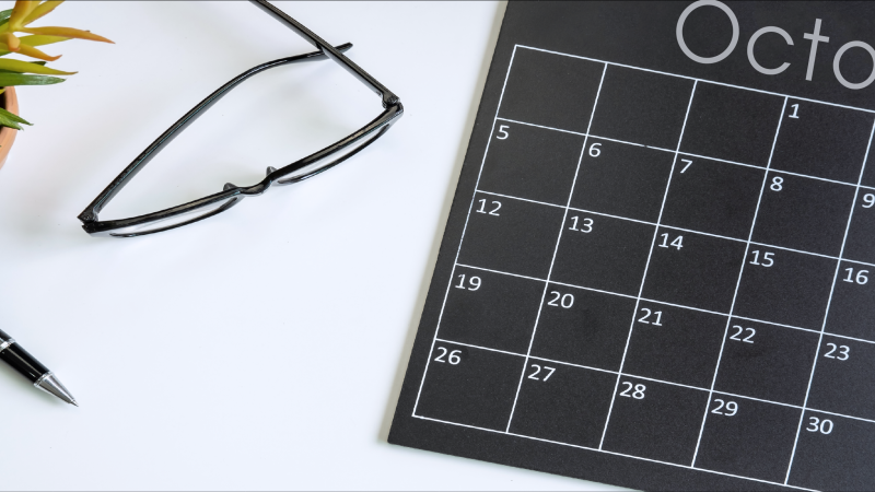 Black and white October calendar sitting on desk next to glasses, a plant, and a pen