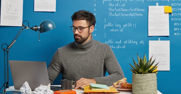 Developer sitting at desk with a laptop and coffee mug in front of him. Lines of code are written on the blue wall behind him and pieces of paper are taped on the wall.